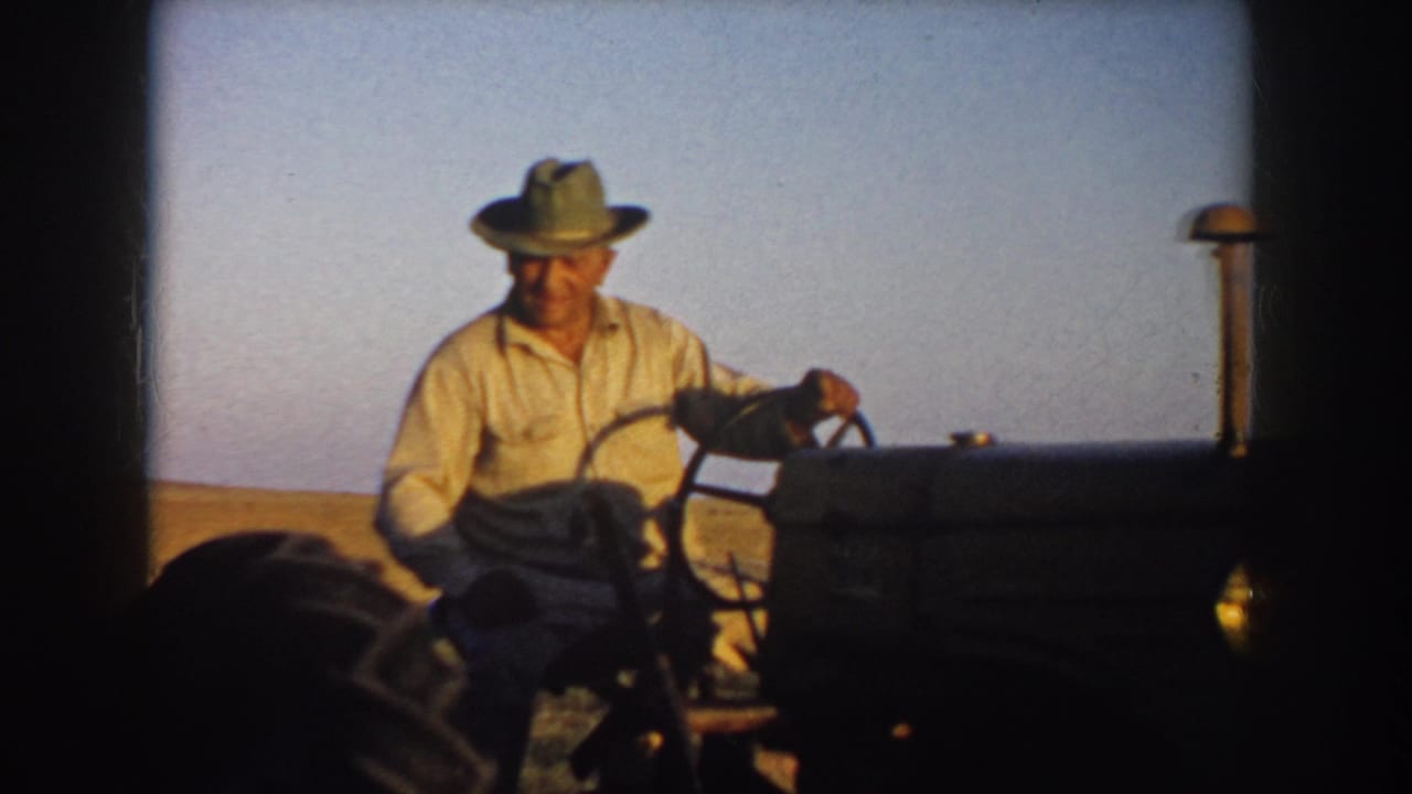 A closer 8mm frame shows the farmer in a light shirt and hat steering the tractor, with the dark hood and open field behind him.