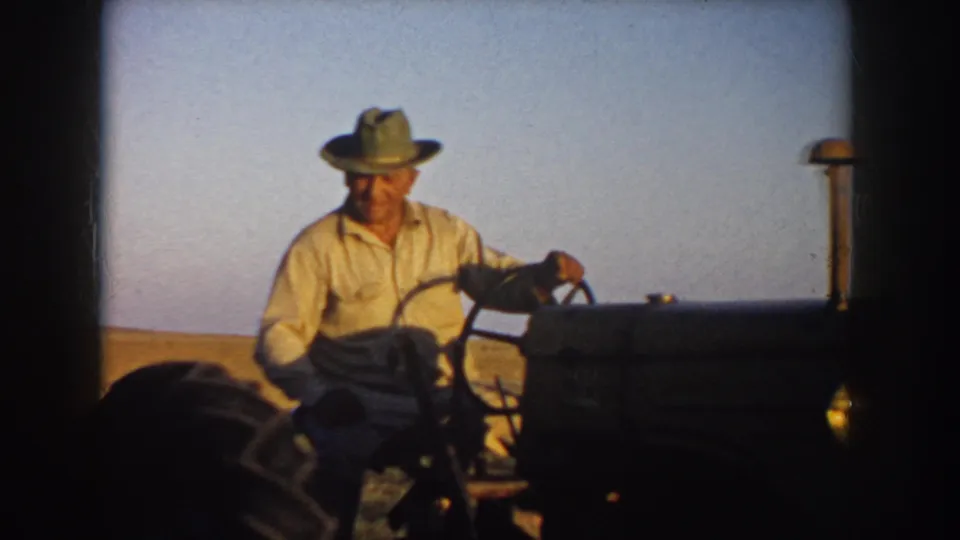 A closer 8mm frame shows the farmer in a light shirt and hat steering the tractor, with the dark hood and open field behind him.