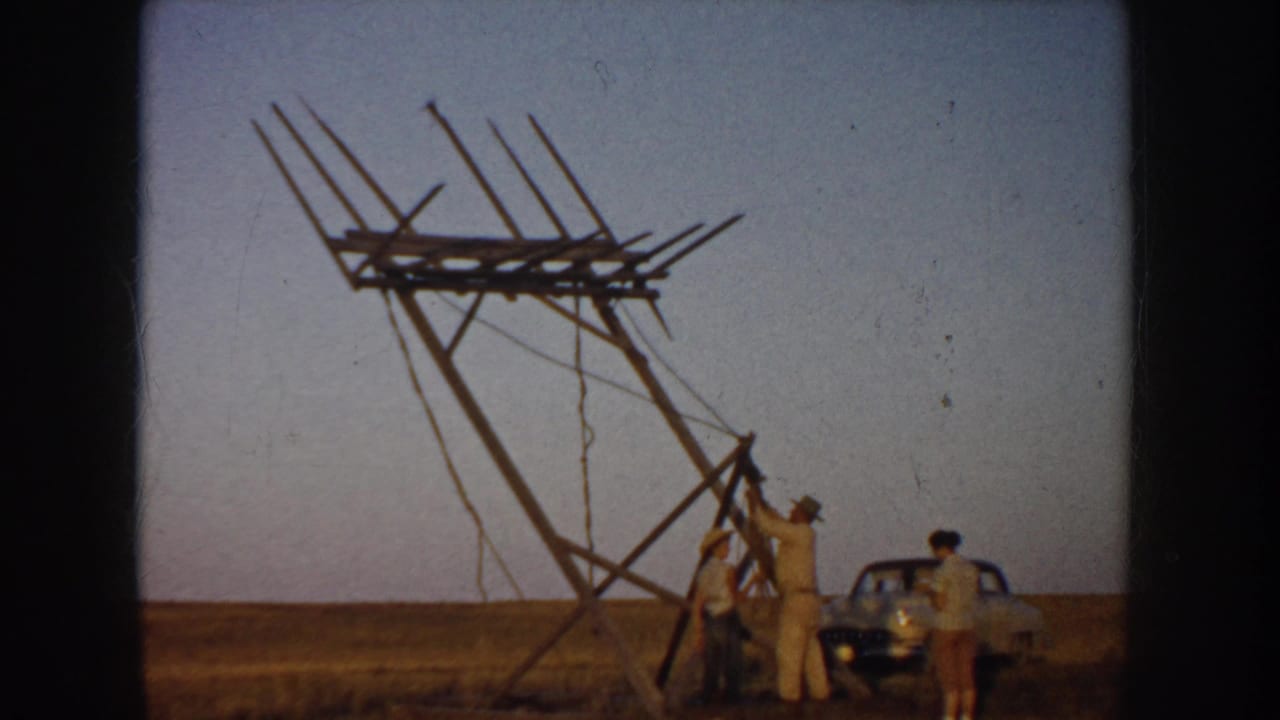 1953 South Dakota home-movie frame showing men assembling a tall wooden antenna-like structure in a flat field beside a parked car.