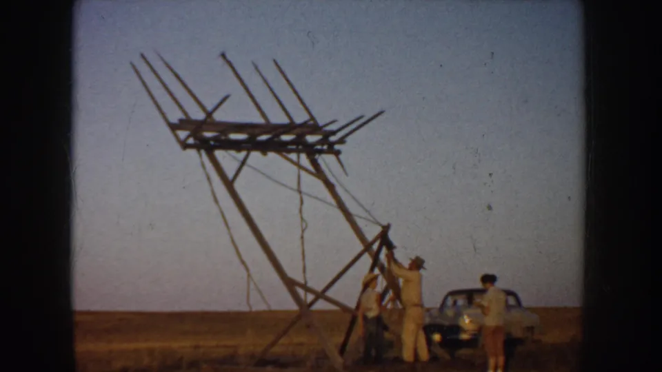 1953 South Dakota home-movie frame showing men assembling a tall wooden antenna-like structure in a flat field beside a parked car.