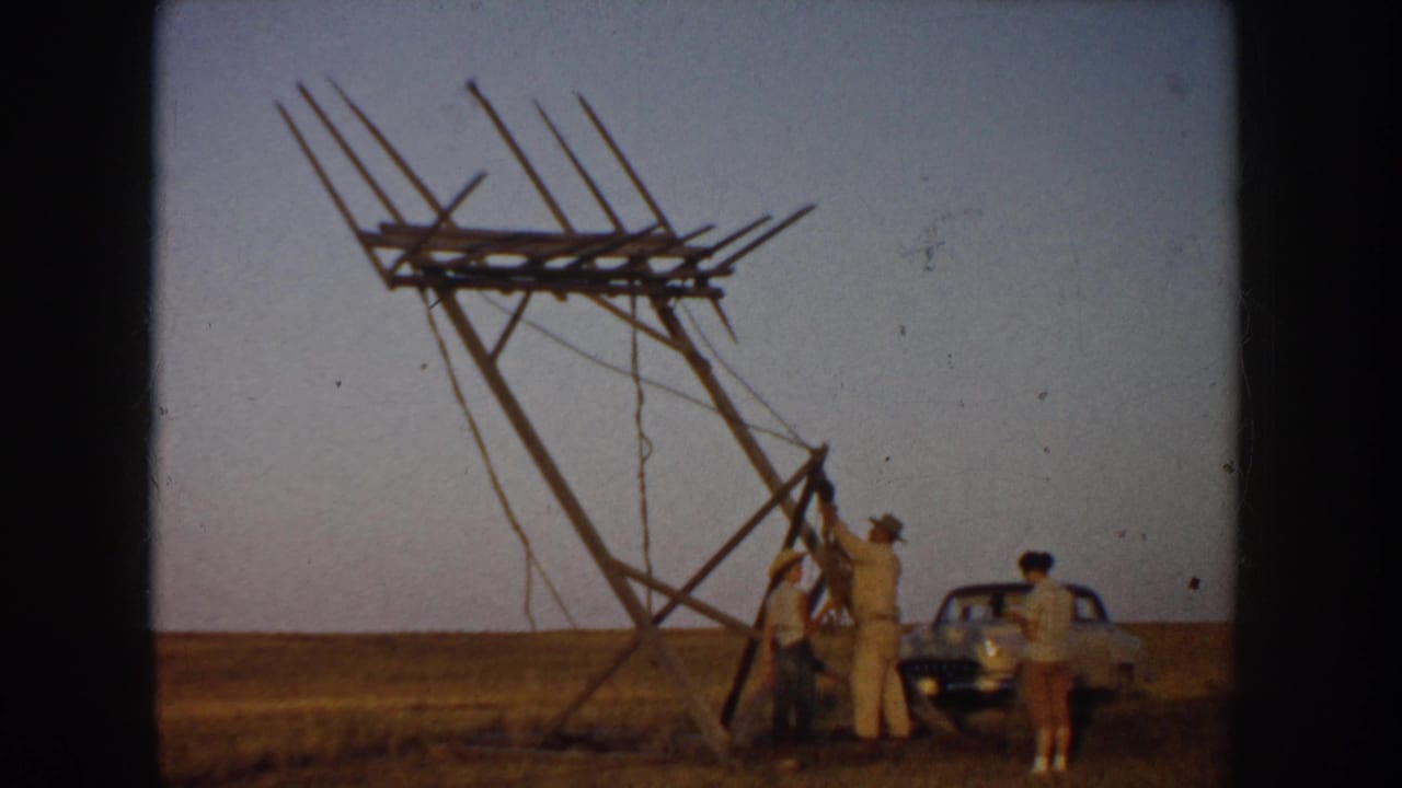 Group of people stand near a tilted wooden frame with a car parked on the right.