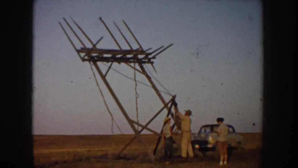 Men appear to adjust or support a wooden antenna structure in a South Dakota field, with a midcentury car and open horizon behind them.