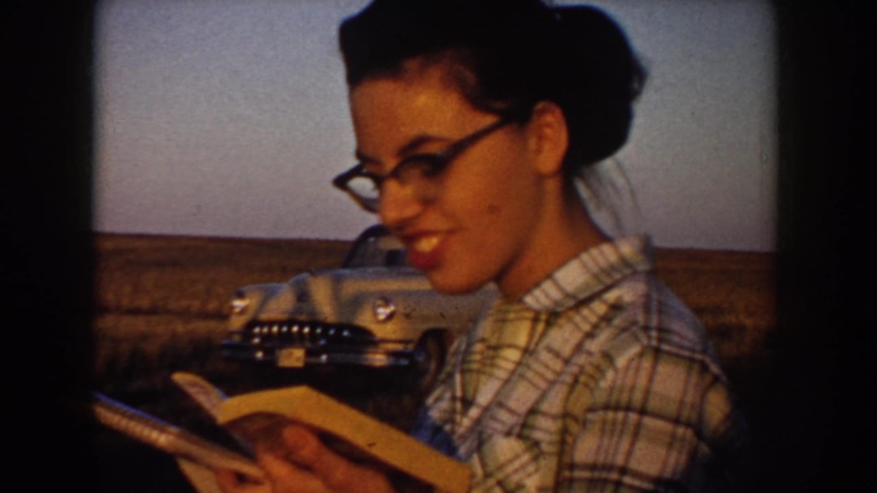 A woman in glasses and a plaid shirt reads and smiles in the foreground, with the rural South Dakota field and 1950s car behind her.