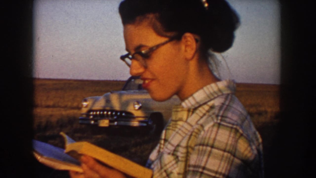 In South Dakota home-movie footage from 1953, a woman in glasses and a plaid shirt reads from a book beside an open field and parked car.