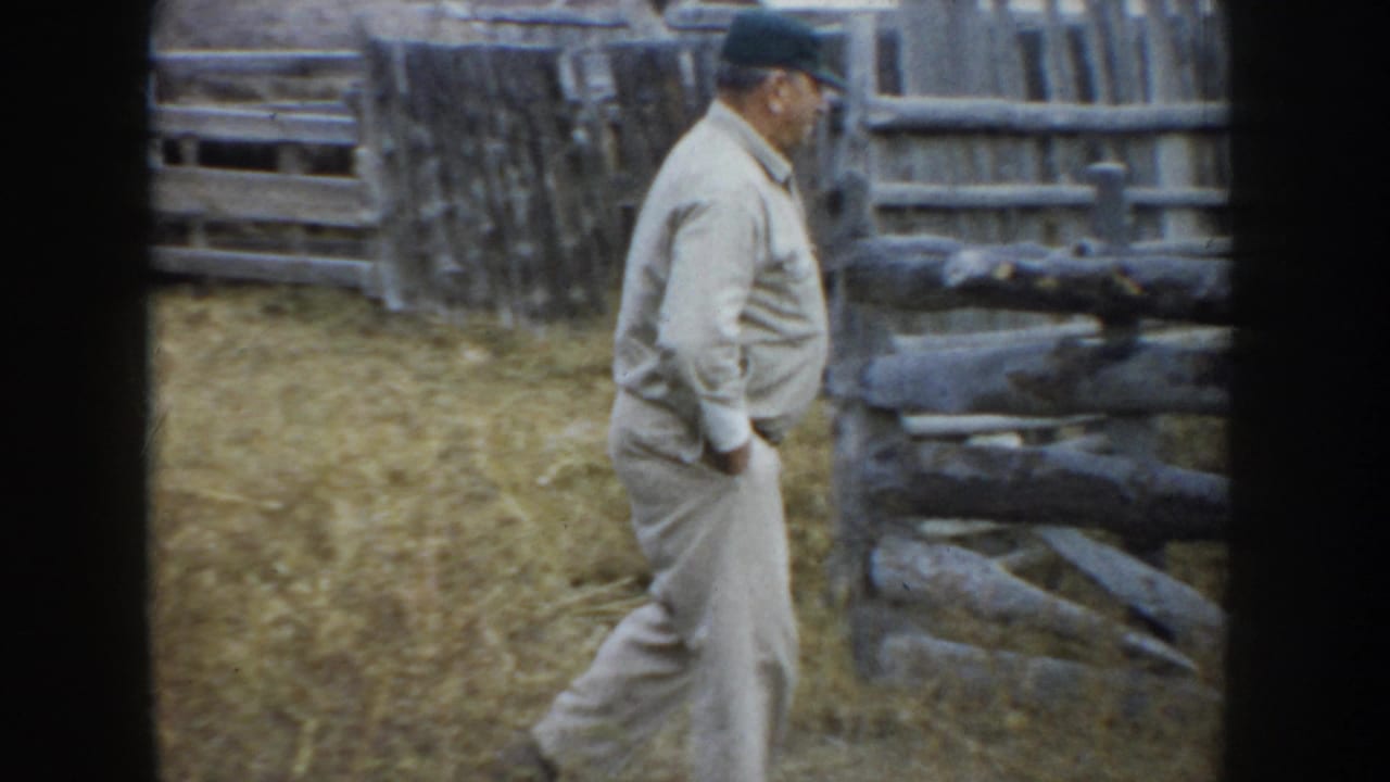 Older man walking beside a weathered wooden fence in dry grass.