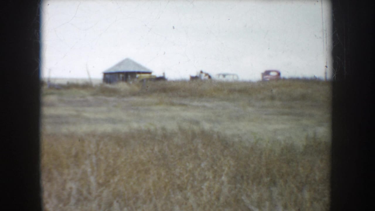 South Dakota ranch field, 1953, with dry grass, distant farm structure, and a small period car on the horizon.