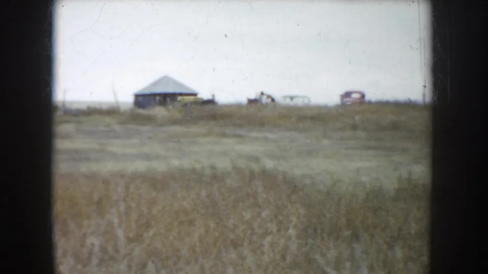 South Dakota ranch field, 1953, with dry grass, distant farm structure, and a small period car on the horizon.