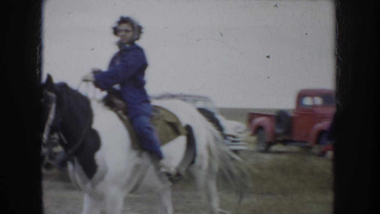 1953 South Dakota home-movie frame showing a woman on a pinto horse with mid-century vehicles nearby.