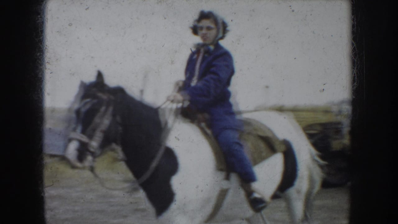 In South Dakota 1953 home-movie footage, a young woman wearing glasses and blue riding clothes sits on a black-and-white pinto horse near farm buildings.