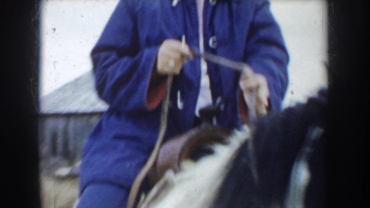 The home-movie frame closely crops the South Dakota rider's blue jacket, hands, reins, saddle, and part of the pinto horse.