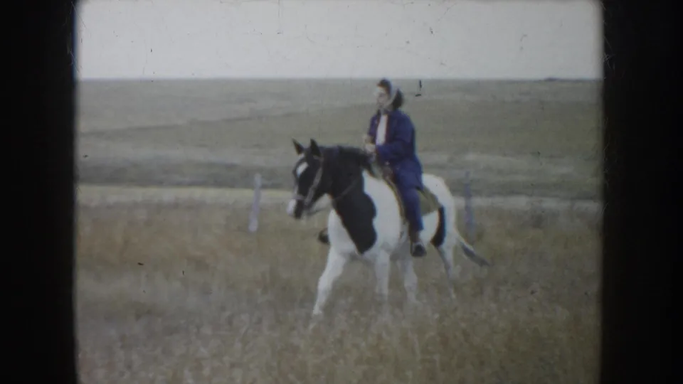 1953 South Dakota frame showing a rider in a dark jacket and hat on a black-and-white horse among dry prairie grass.