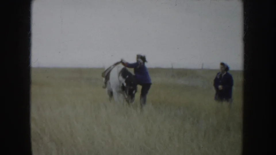 Two people in dark clothing handle a white horse on the South Dakota prairie, with tall grass and a flat horizon behind them.