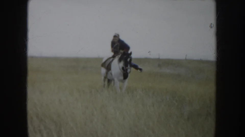A rider sits on a white horse in tall South Dakota prairie grass, filmed from a distance in soft 8mm color.