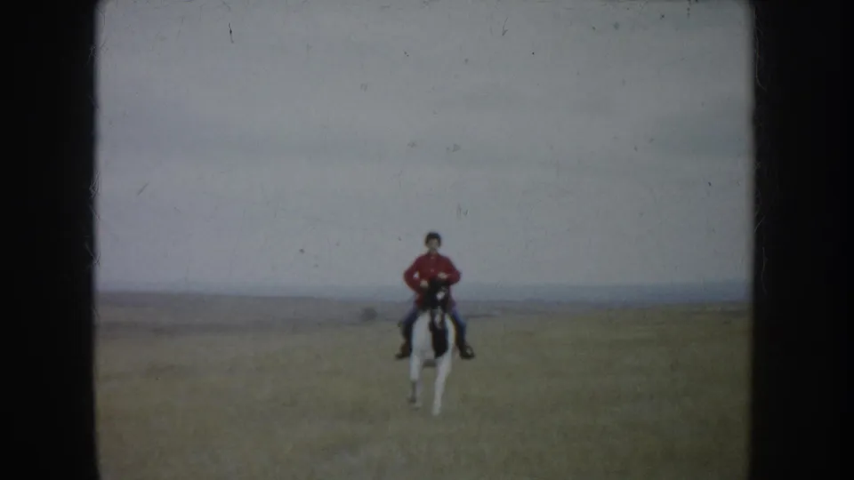 In 1953 South Dakota footage, a rider in a red shirt approaches across an open field on a white-and-dark horse.