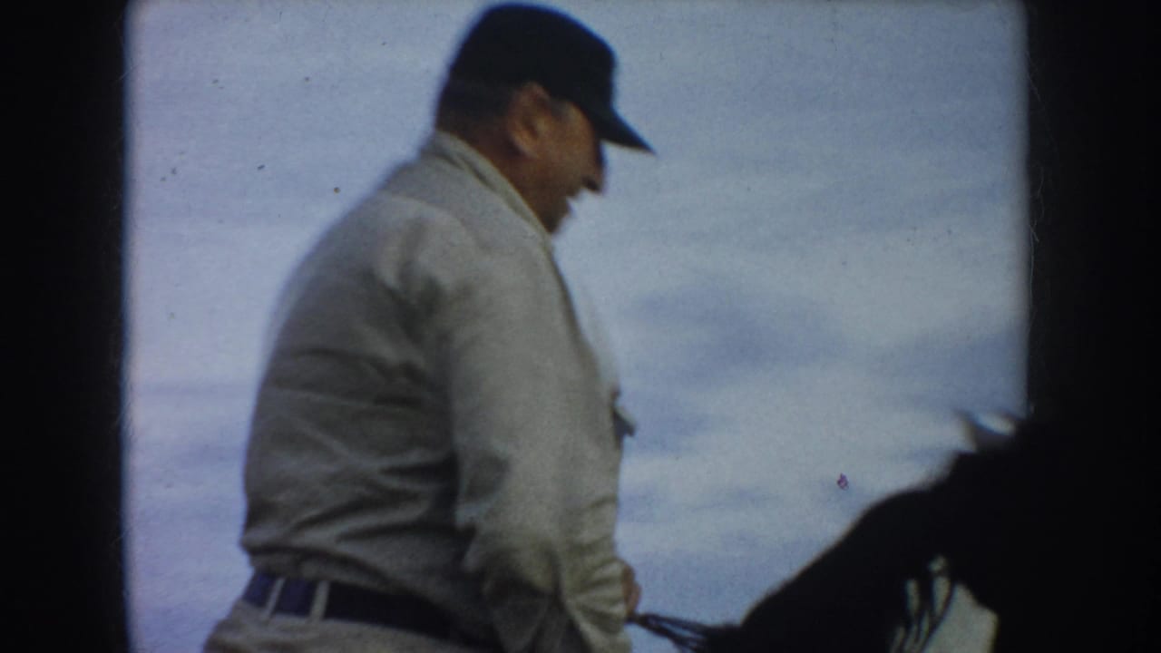 A second rider wearing a light jacket and dark cap passes close to the camera beside a dark horse.