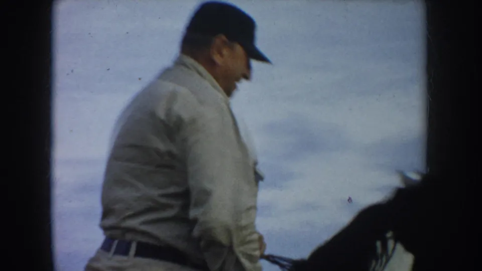 A second rider wearing a light jacket and dark cap passes close to the camera beside a dark horse.