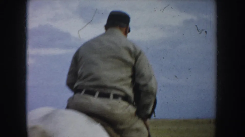 In 1953 South Dakota home-movie footage, a man in light uniform-style clothing rides a spotted horse away across prairie grass.