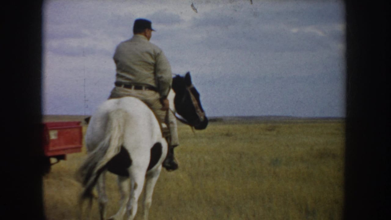 The man rides farther away across the South Dakota grassland on a white-and-dark-spotted horse, with a red object partly visible behind him.