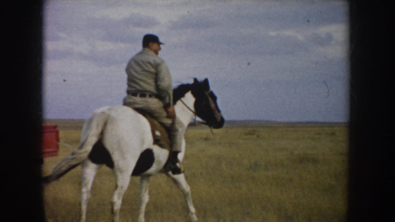 1953 South Dakota home-movie frame of an adult man on a white-and-black horse walking through open prairie grass.