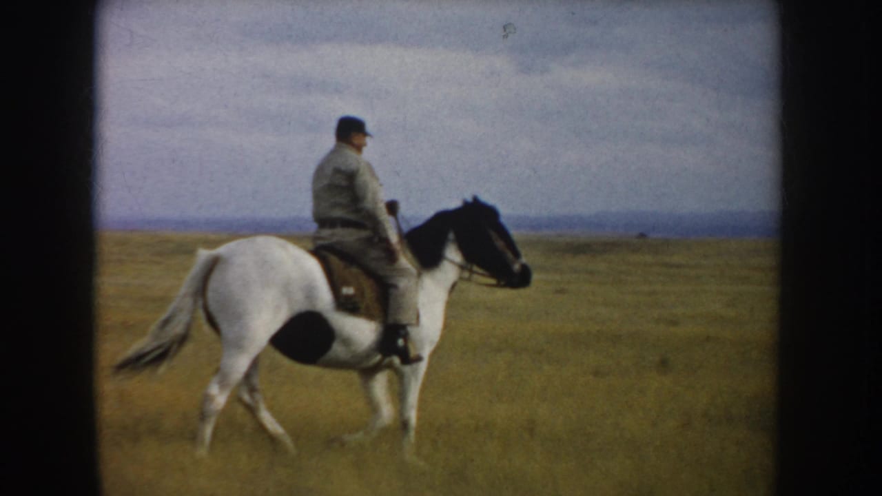Side-on 1953 home-movie view of a man riding a white horse with black patches through South Dakota prairie.