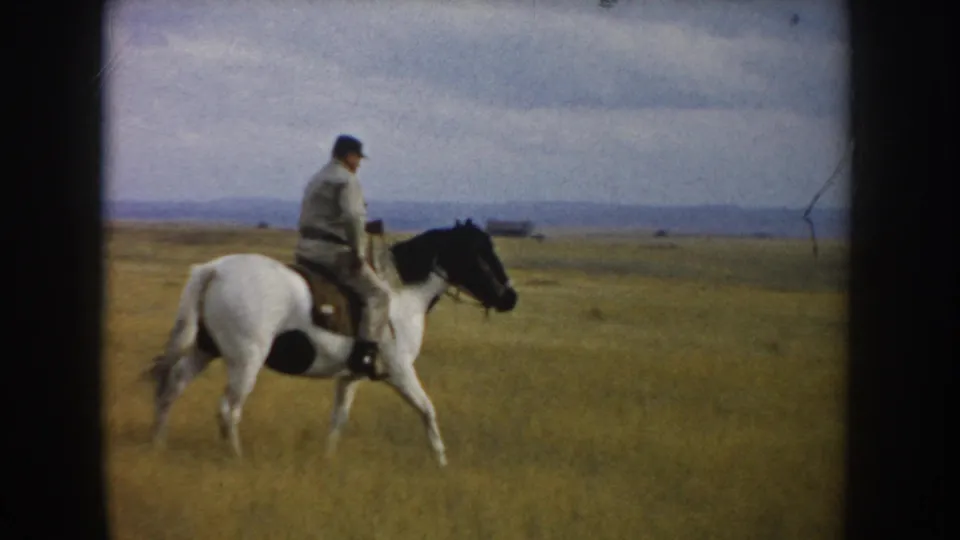 Adult rider on a white-and-black horse crosses open South Dakota grassland with a distant low horizon behind them.