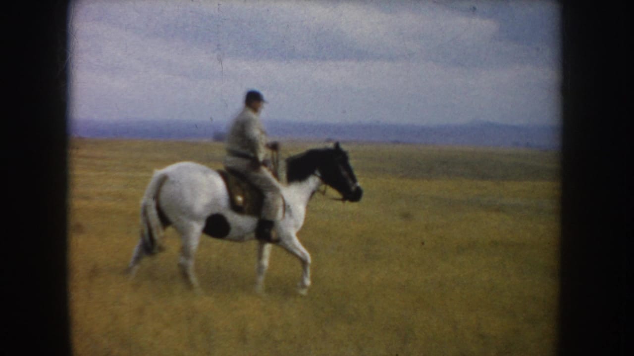 In 1953 South Dakota footage, an adult rider on a white horse with black markings moves through open prairie under a pale sky.