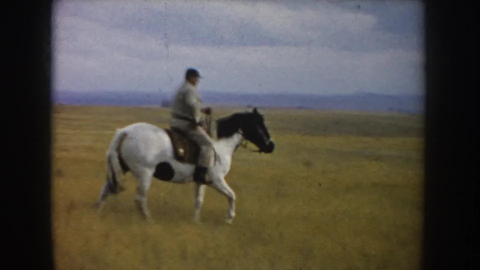 In 1953 South Dakota footage, an adult rider on a white horse with black markings moves through open prairie under a pale sky.