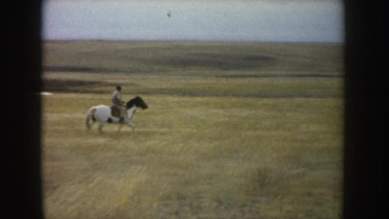 The rider continues across South Dakota prairie on a white-and-dark horse, framed small against a broad grassland horizon.
