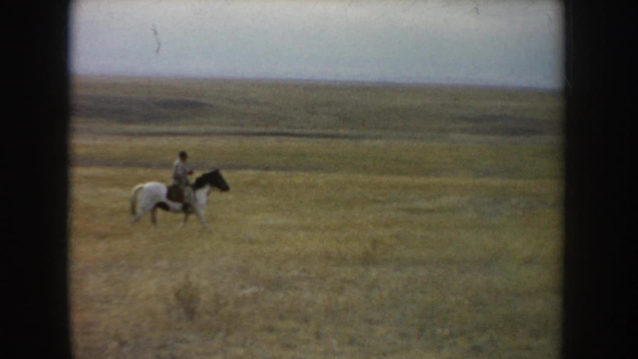 A distant adult rider on a marked horse moves through South Dakota grassland, with low rolling terrain visible on the horizon.