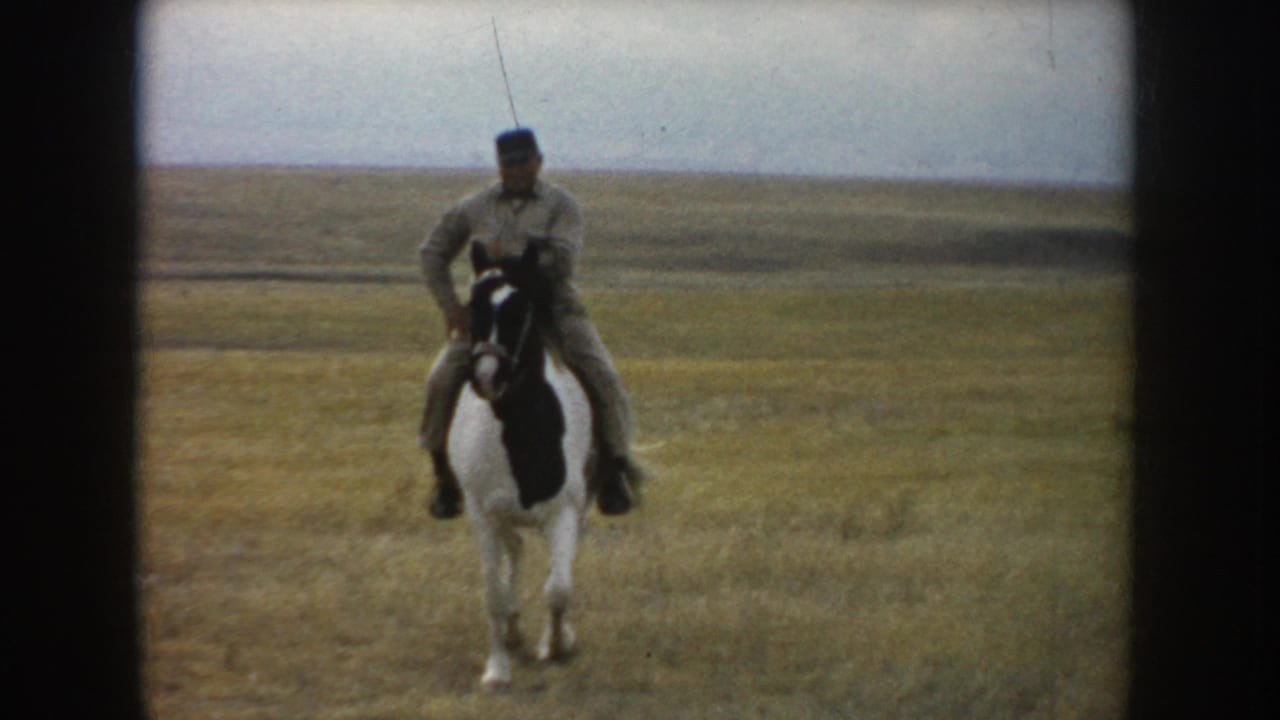In South Dakota home-movie footage from 1953, an adult rider on a pinto horse faces the camera in an open field under a pale sky.