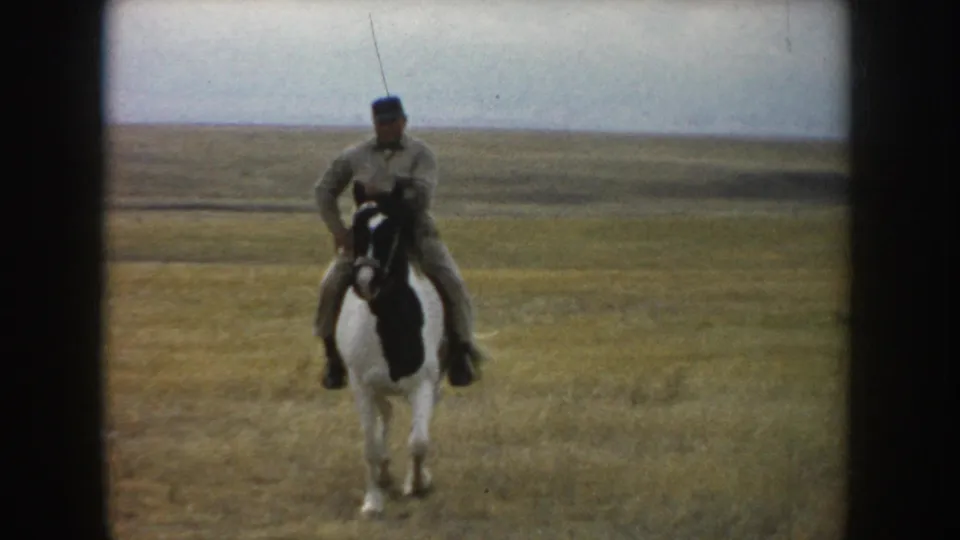 In South Dakota home-movie footage from 1953, an adult rider on a pinto horse faces the camera in an open field under a pale sky.