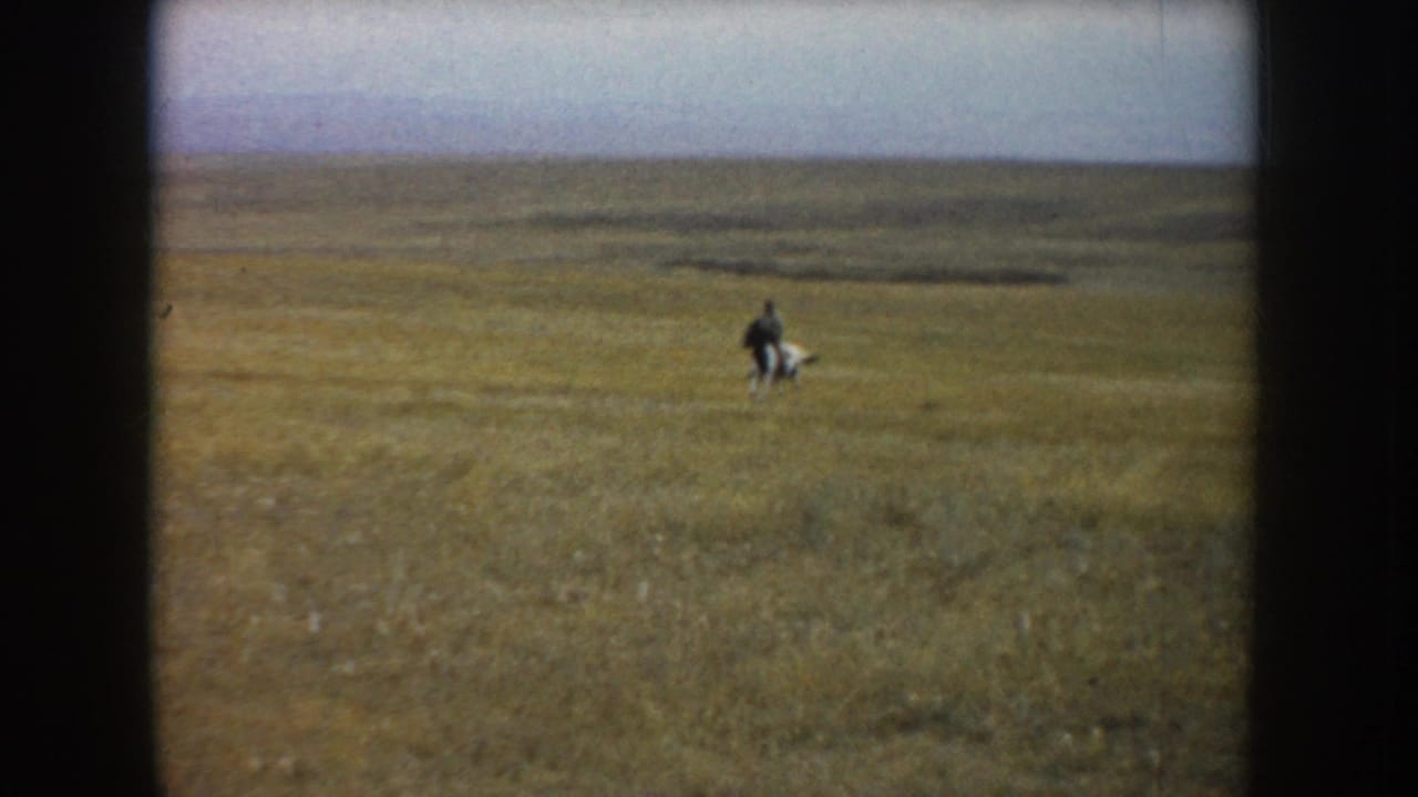 In 1953 South Dakota home-movie footage, a distant man rides a black-and-white horse across broad prairie under a pale sky.