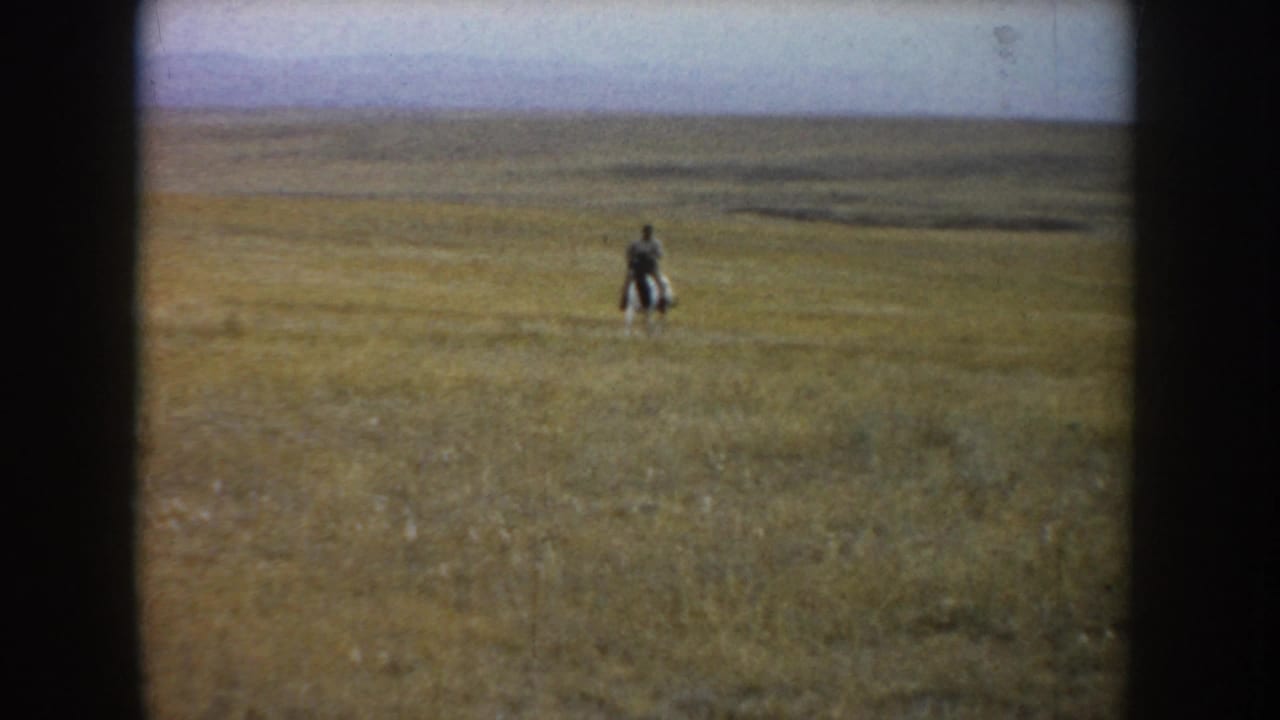 Soft 8mm footage shows the rider and black-and-white horse farther out in the grassland, with the prairie stretching behind them.