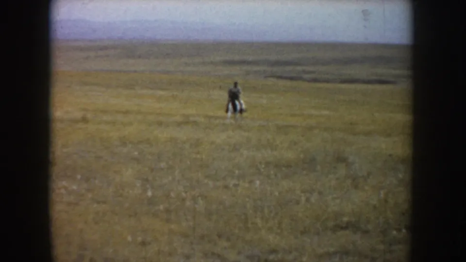 Soft 8mm footage shows the rider and black-and-white horse farther out in the grassland, with the prairie stretching behind them.