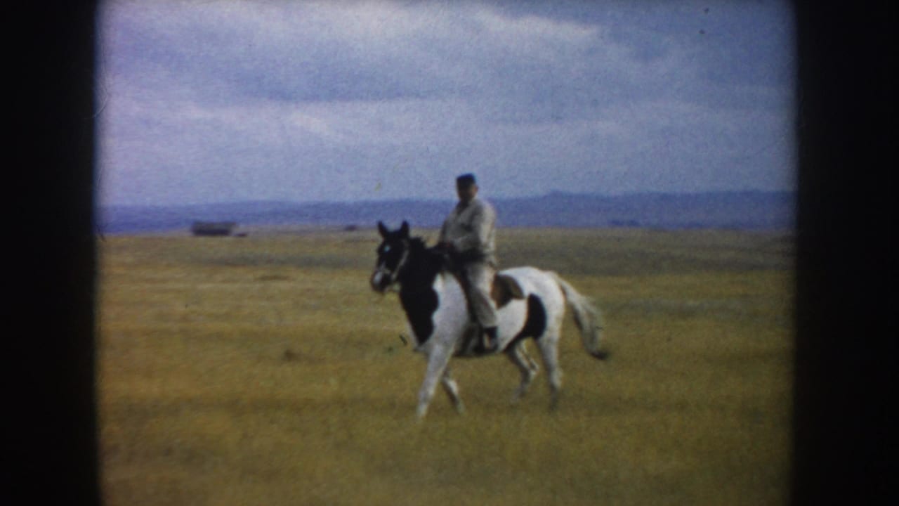 Man riding a black-and-white pinto horse across an open grassy field.