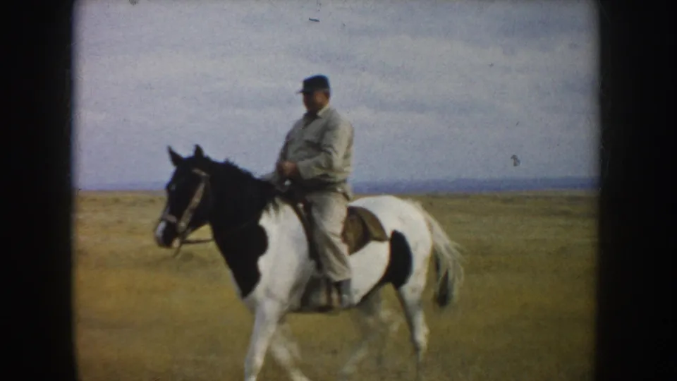 1953 South Dakota 8mm frame of a man in light clothing riding a pinto horse across an open grassy plain.
