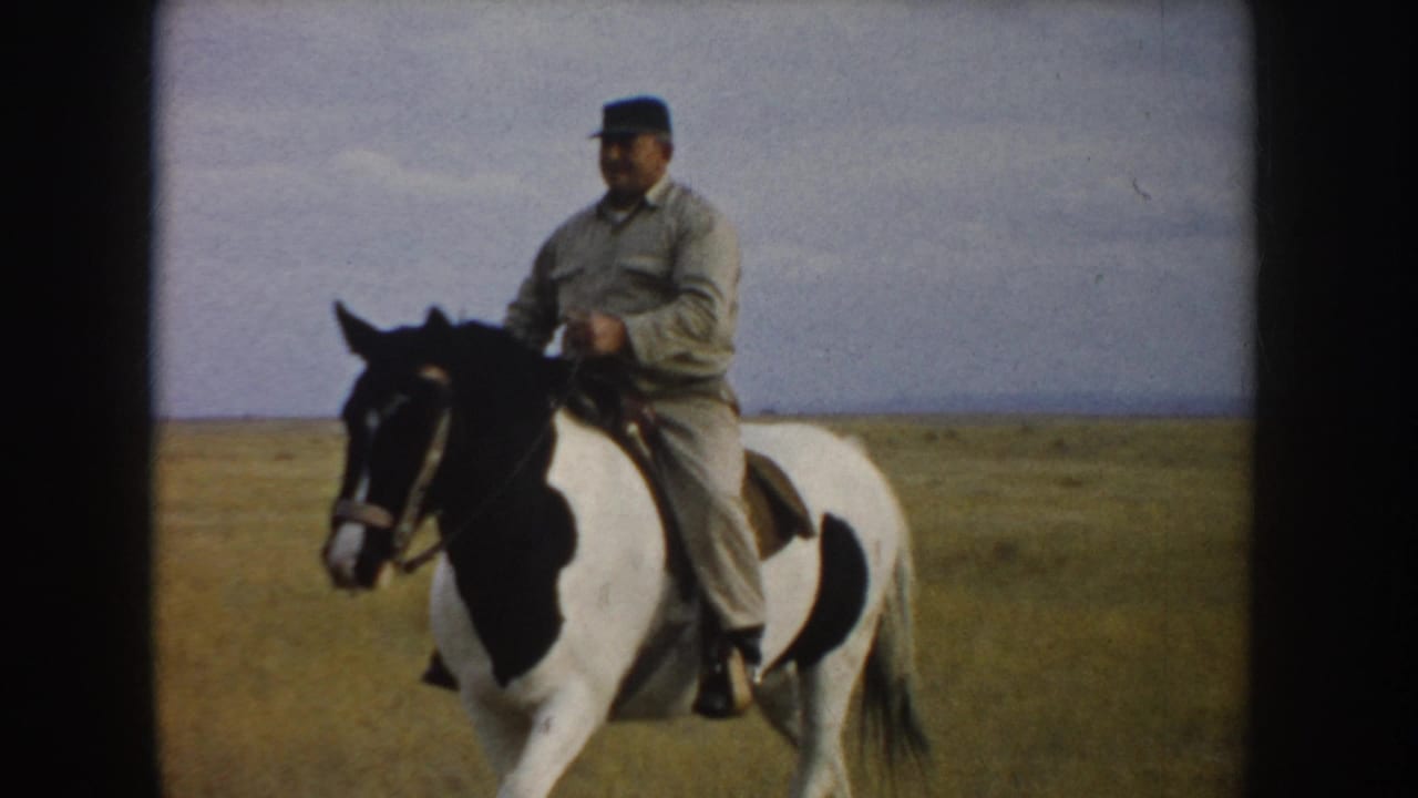 A man in light clothing and a dark cap rides a black-and-white pinto horse through South Dakota grassland in 1953 footage.