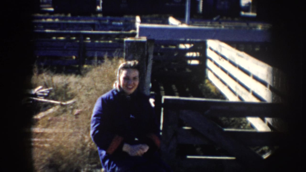 Girl smiles toward the camera beside wooden pens in a South Dakota livestock-market home movie.