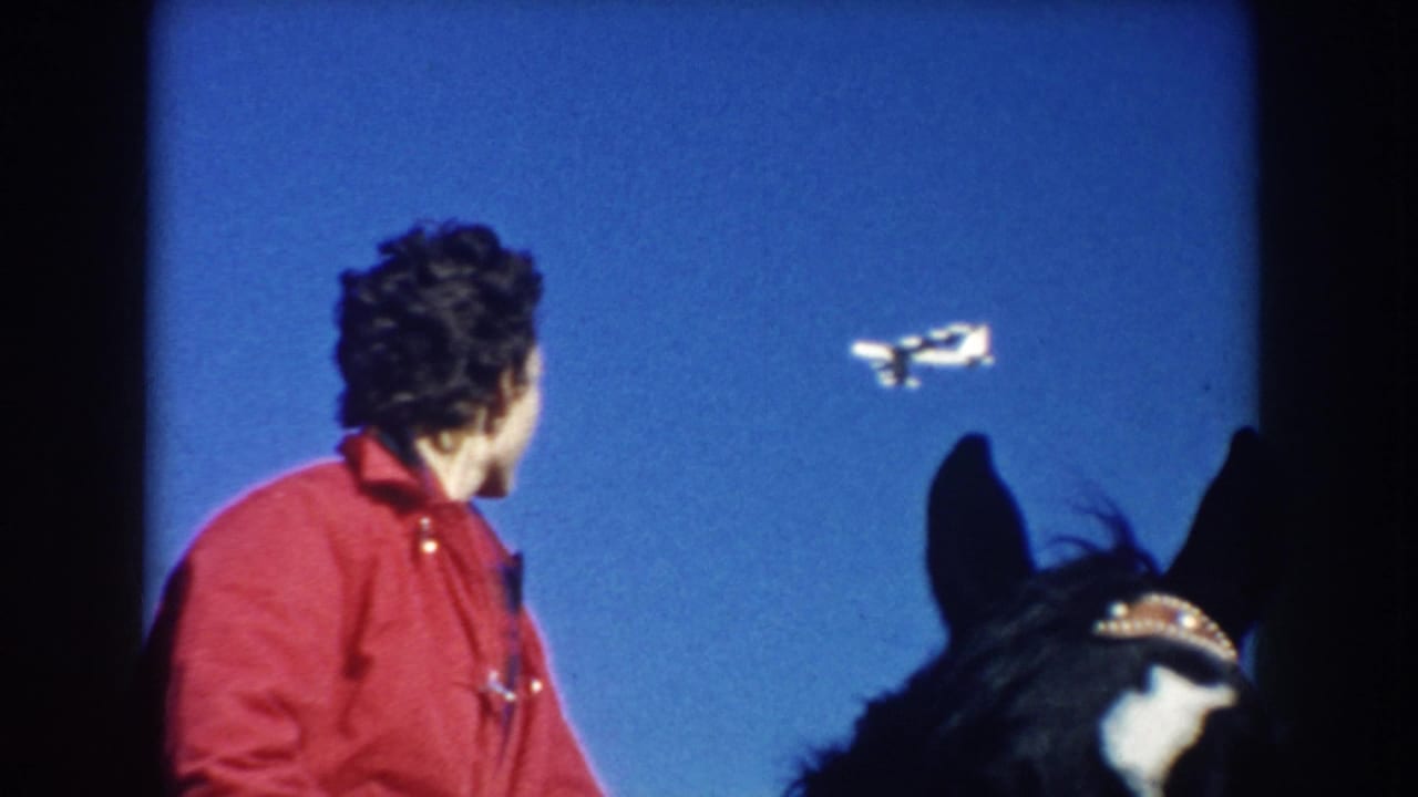 1953 South Dakota frame showing an adult rider in a red jacket on a dark horse watching a small airplane pass against a clear blue sky.