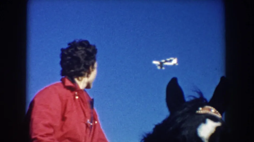 1953 South Dakota frame showing an adult rider in a red jacket on a dark horse watching a small airplane pass against a clear blue sky.