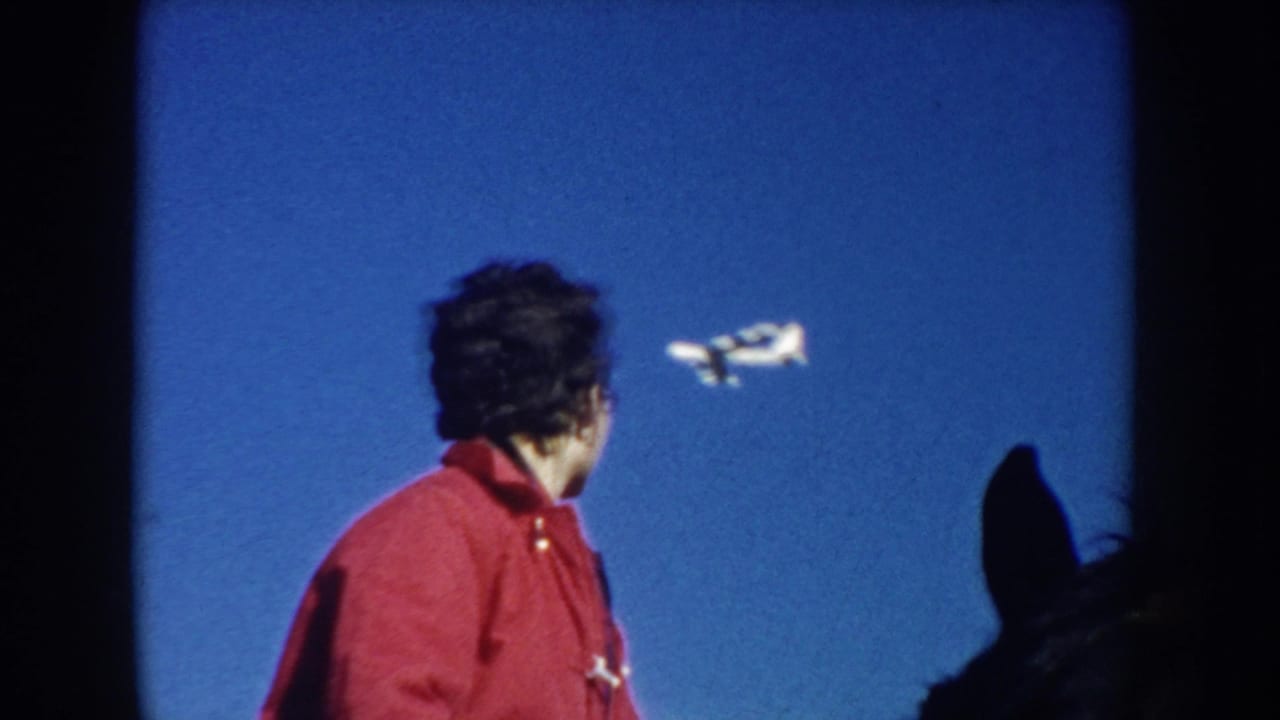 In 1953 South Dakota home-movie footage, a woman in a red coat looks toward a small airplane passing overhead against a clear blue sky.