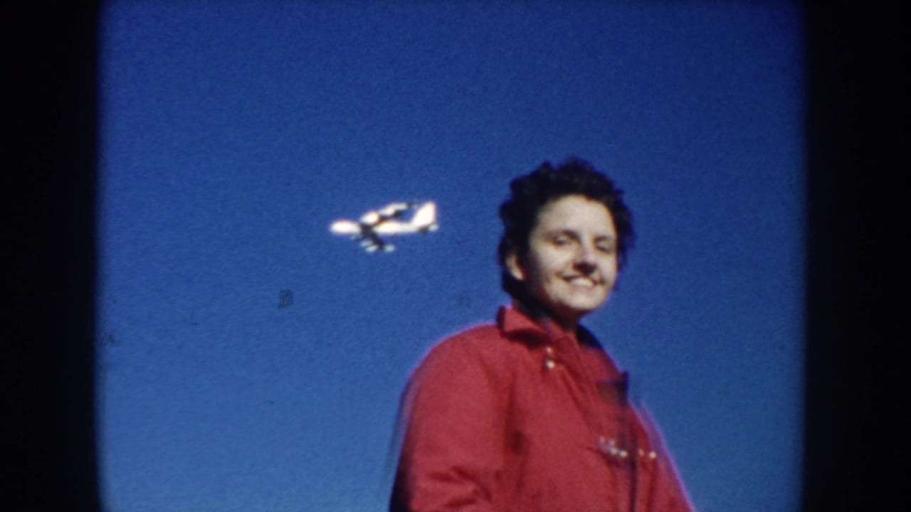 Woman in a red coat faces the camera as a small airplane passes behind her.