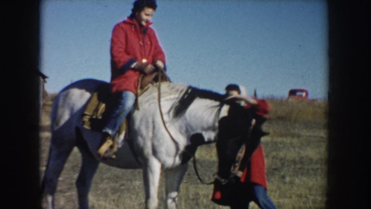 The woman in the red coat rides a white horse through an open South Dakota field, with another person partly visible near the horse.