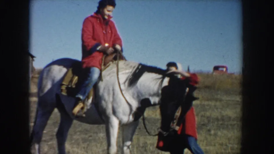The woman in the red coat rides a white horse through an open South Dakota field, with another person partly visible near the horse.