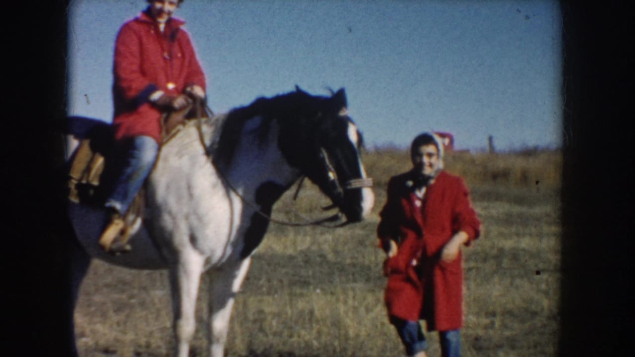1953 South Dakota home-movie frame showing a man riding a pinto horse as a woman in a red coat stands nearby in dry pasture.