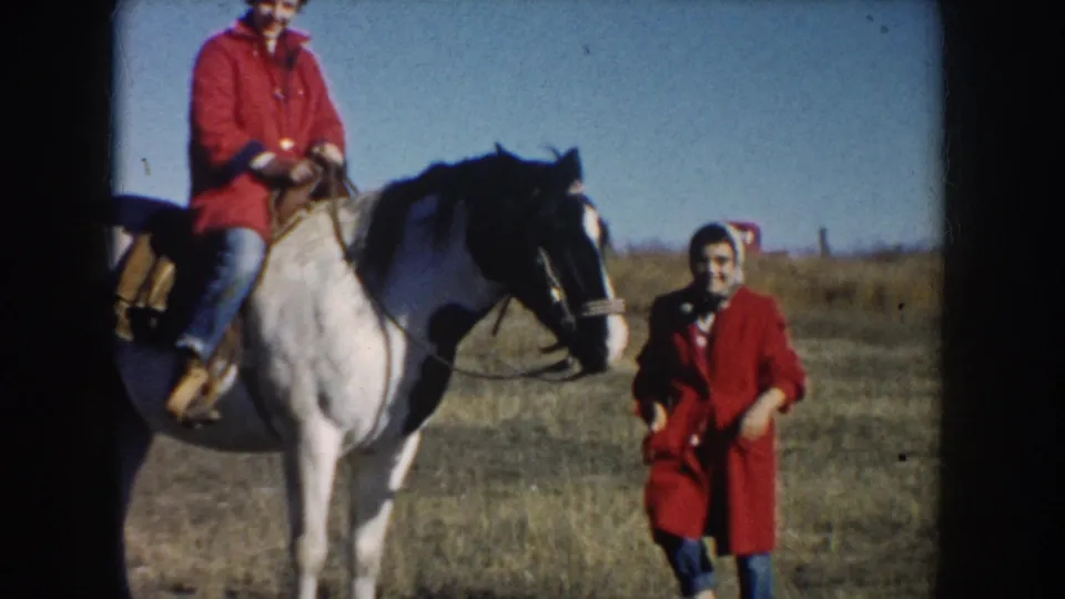 1953 South Dakota home-movie frame showing a man riding a pinto horse as a woman in a red coat stands nearby in dry pasture.