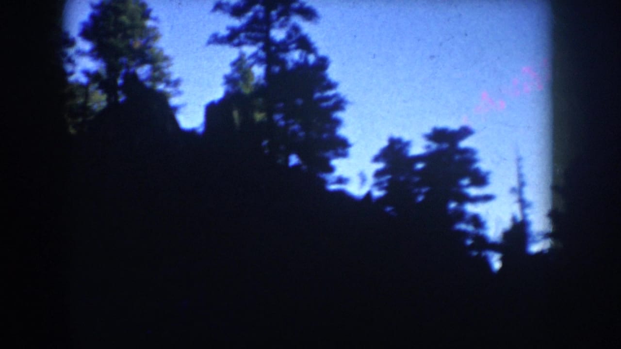 1953 South Dakota 8mm frame with dark pine silhouettes, blue sky, and a partly visible rocky hillside.
