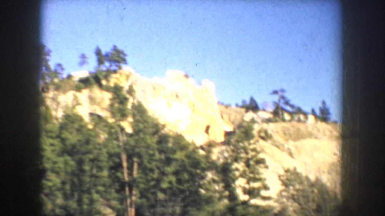 1953 South Dakota 8mm view of a pale rocky cliff rising above evergreen trees under clear blue sky.