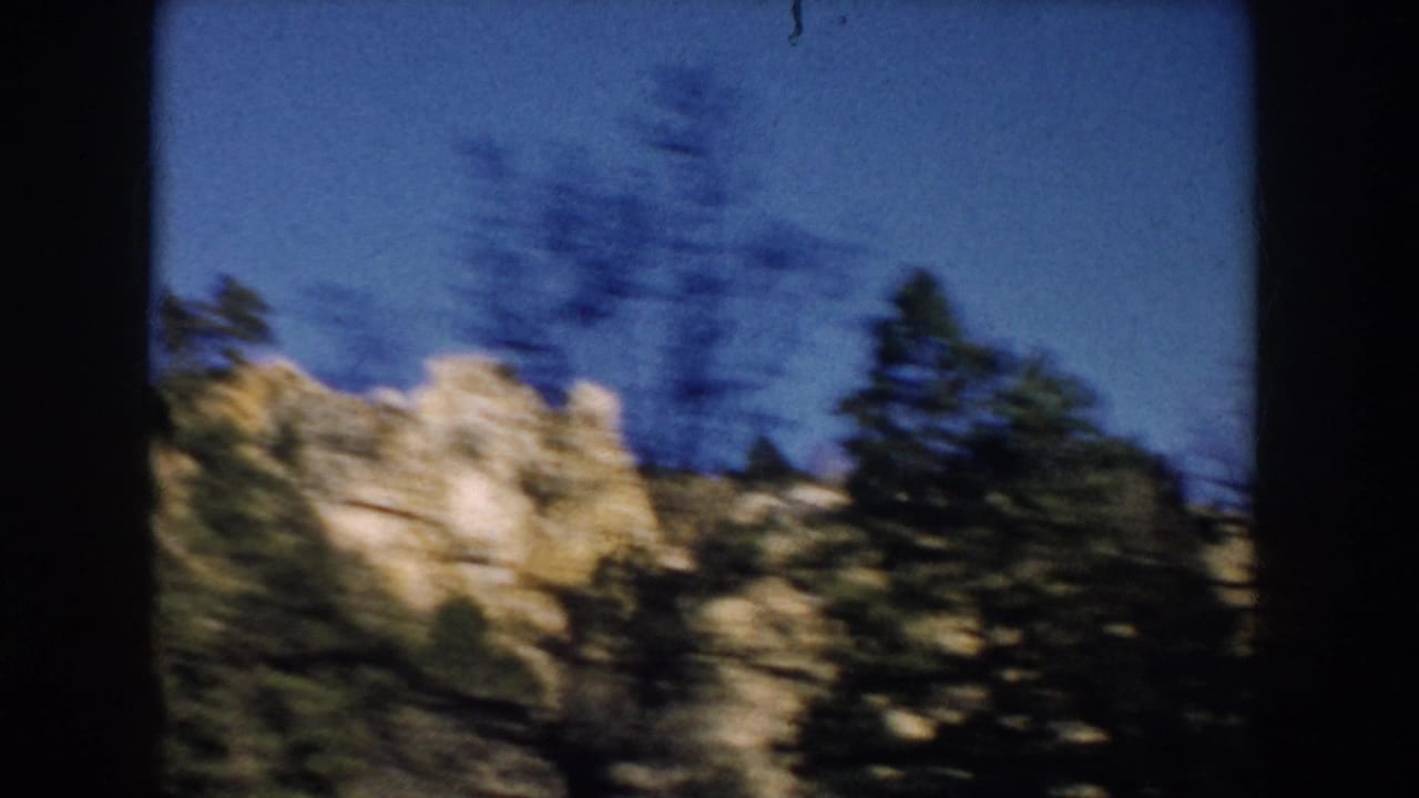1953 South Dakota 8mm frame with motion-blurred evergreens and pale rocky formations under blue sky.
