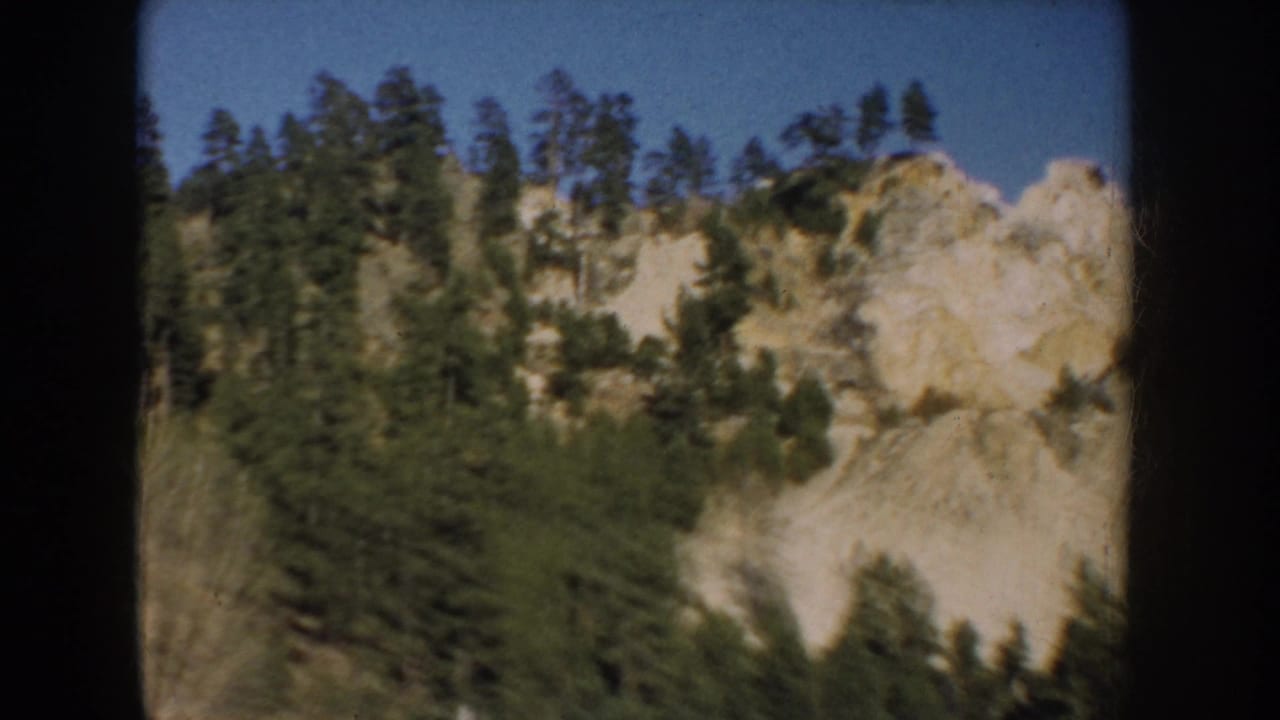 1953 South Dakota 8mm frame showing a pine-covered rocky slope with pale cliff sections below blue sky.